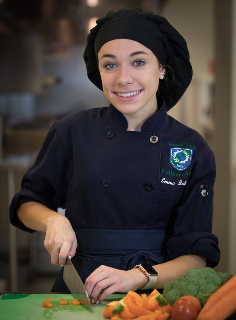 A student in full chef's regalia cuts veggies.