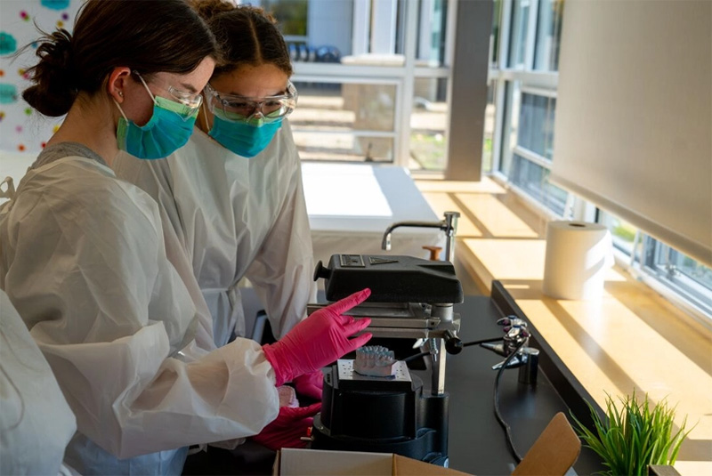 Two students in full protection gear, work at a microscope.
